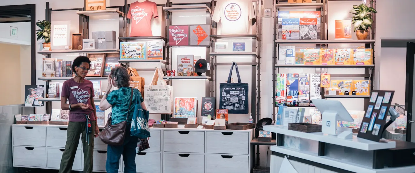 Two women browsing framed art and books in a bright shop interior.