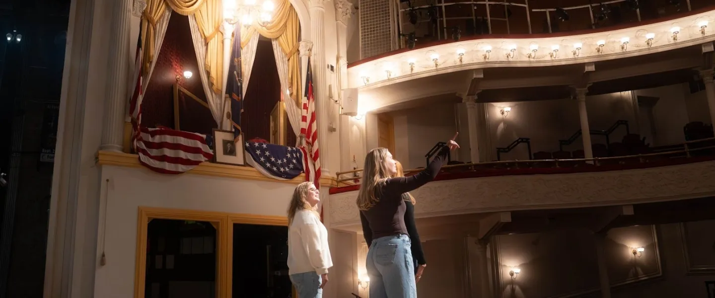 Two performers on stage beneath ornate theater balconies and chandelier.
