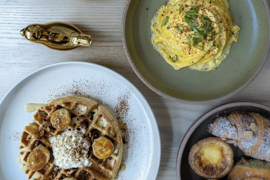 A top-down view of a breakfast spread, featuring a plate of waffles topped with whipped cream and banana slices, a dish of scrambled eggs garnished with green onions, and a bowl of assorted pastries.