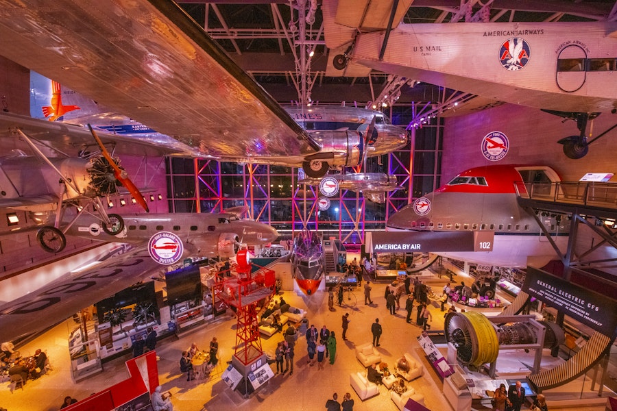 An aerial view of the "America by Air" exhibit at the National Air and Space Museum, showcasing a dynamic collection of aircraft illuminated by vibrant lighting.