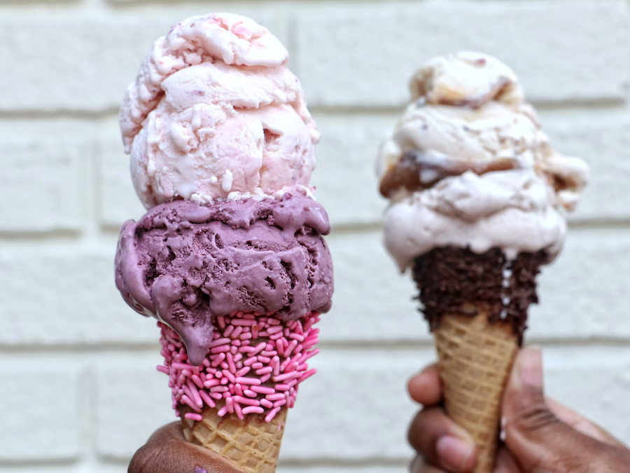 Two hands holding ice cream cones topped with double scoops. The left cone features pink and purple ice cream with pink sprinkles, while the right cone has scoops of brown and white ice cream with chocolate sprinkles, against a white brick background.