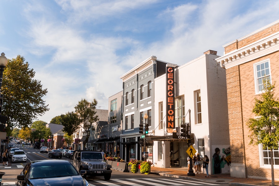 Pedestrians and cars pass by storefronts under a bright Georgetown sign on a bustling street in the historic Georgetown neighborhood of Washington, DC.