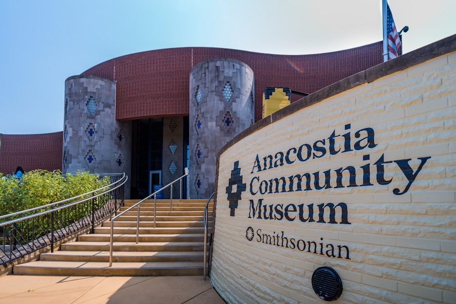 Exterior view of the Smithsonian’s Anacostia Community Museum, featuring distinctive cylindrical architectural elements and a curved yellow brick wall with the museum’s name and logo. An American flag flies at half-mast above the building.