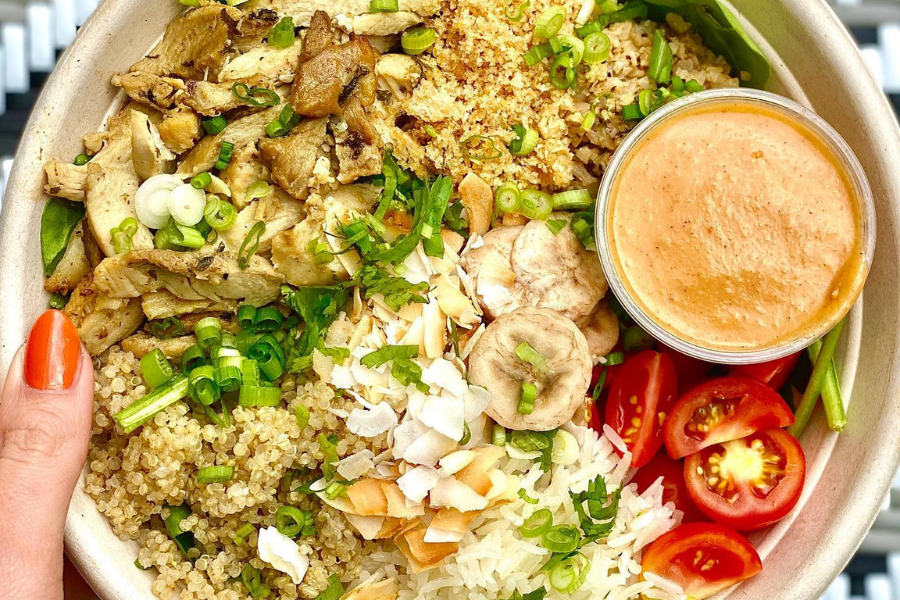 A colorful grain bowl from Immigrant Food in Washington, DC, featuring grilled chicken, quinoa, white rice, cherry tomatoes, coconut flakes, and a creamy dipping sauce.