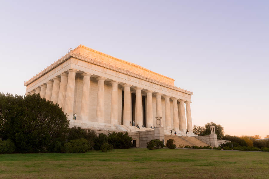 The Lincoln Memorial in Washington, DC, glows with warm sunset light as visitors walk along its steps and surrounding lawn.