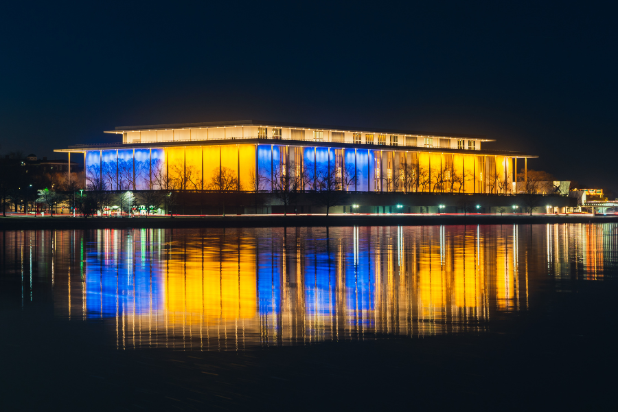 The Kennedy Center in Washington, DC glows in vivid blue and yellow lights, reflecting off the Potomac River on a clear night.