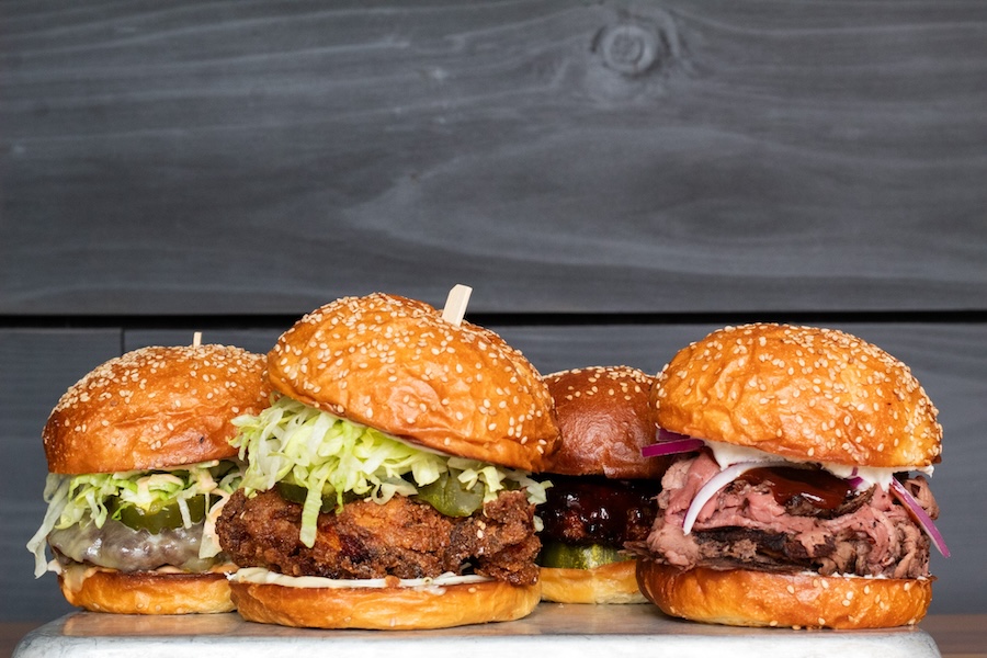 A close-up of four gourmet sandwiches from Shilling Canning Company, layered with meats, pickles, slaw and served on sesame buns.