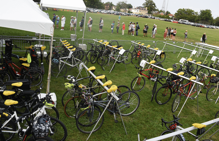 Rows of parked bicycles wait in a fenced valet area as people line up nearby.