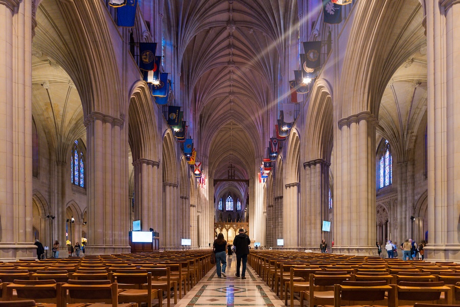Visitors walk down the nave of Washington National Cathedral beneath soaring Gothic arches and flags.