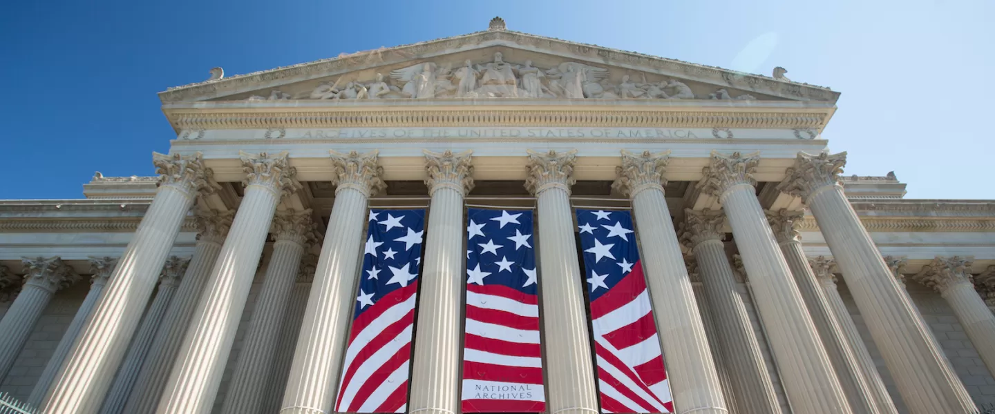The grand neoclassical facade of the National Archives features towering columns and large banners with the American flag.