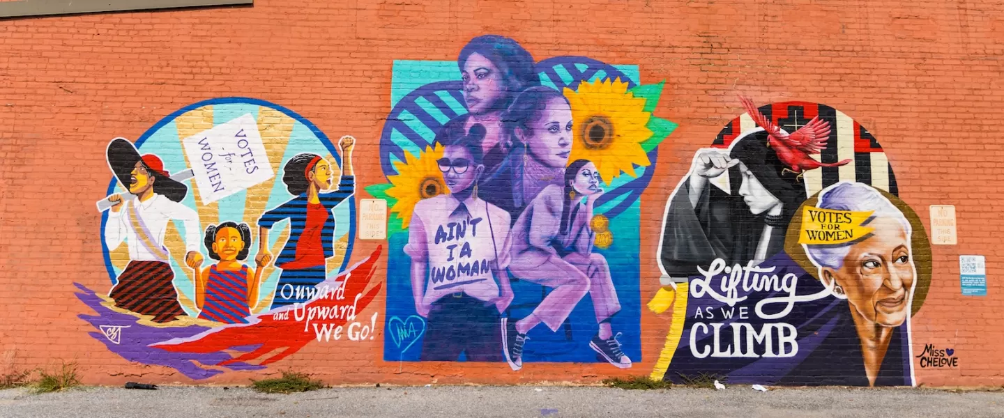 Colorful mural on a red brick wall in Anacostia celebrating women’s suffrage and empowerment. The artwork features historical and modern women of color, sunflowers, protest signs with messages like “Votes for Women” and “Ain’t I a Woman."