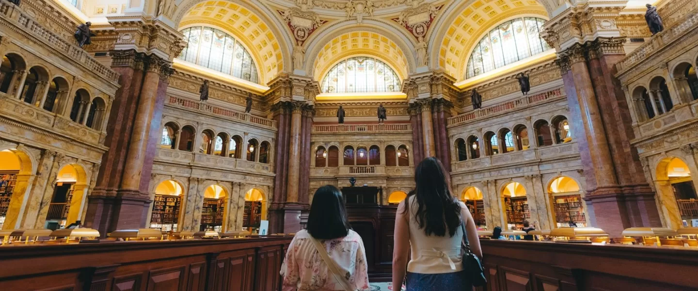 Two people gaze out into the Library of Congress' Reading Room, with grand arches and architecture. 