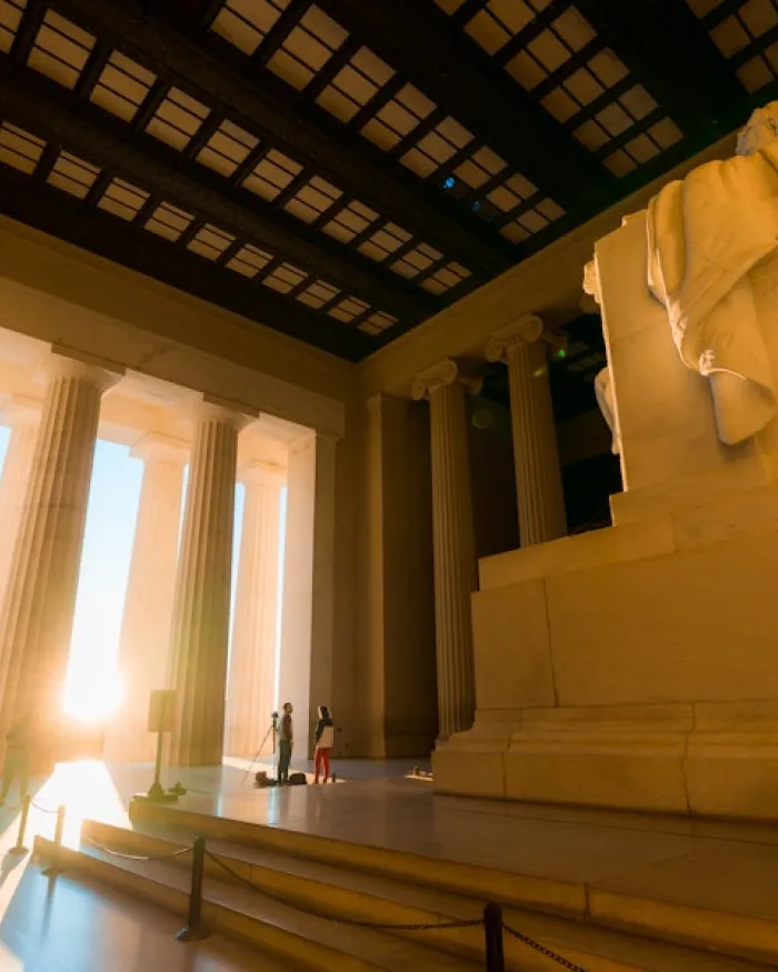 Warm sunlight pours through the columns of the Lincoln Memorial as visitors stand near the towering statue inside the historic monument.